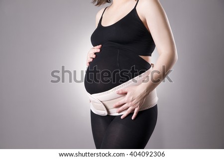 Young caucasian pregnant woman with orthopedic support belt. Studio shot on a gray background