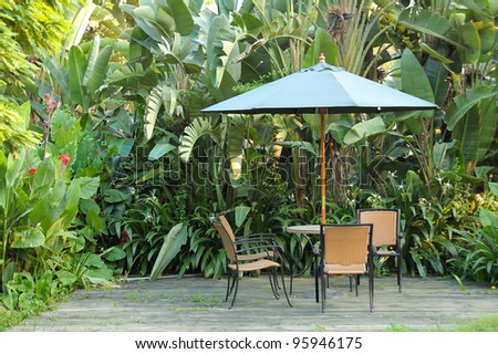 Garden furniture - rattan chairs and table under umbrella on a wooden floor by the banana trees background at garden