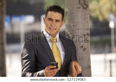 Businessman relaxing next to a tree at the city park, sending short messages over his cellphone