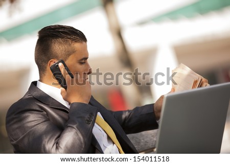 Businessman seated in a bench park, working with his laptop, calling at the cellular and reading his newspaper