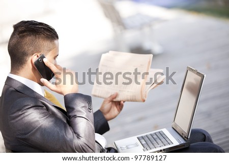 Businessman sited in a bench working with his laptop, calling at the cellular and reading his newspaper
