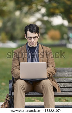 Young Man Sitting On Park Bench, Working On His Laptop