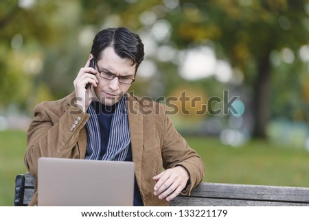 Young Man Sitting On Park Bench, Multi-tasking With Mobile Phone And Laptop Computer