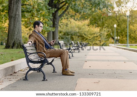 Young Man Reading With A Digital Tablet On A Park Bench