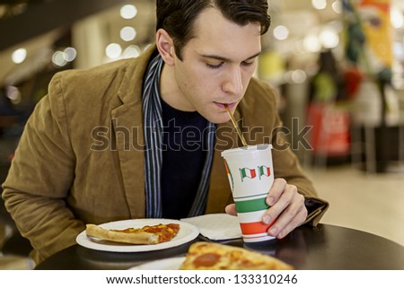 Happy Young Man Eating Pizza At The Food Court In A Mall