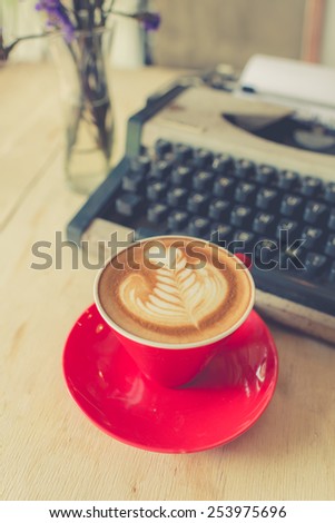 Red latte coffee cup with purple flower and typewriter on wooden background.