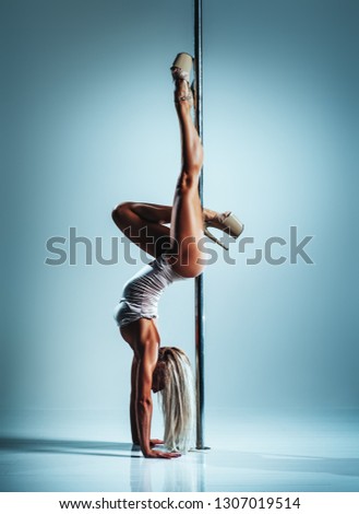 Young slim sexy blond woman in white clothing pole dancing upside down on white wall background