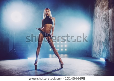 Young slim sexy blond woman dancing in big hall interior with smoke and blue lights. Tattoo on body.