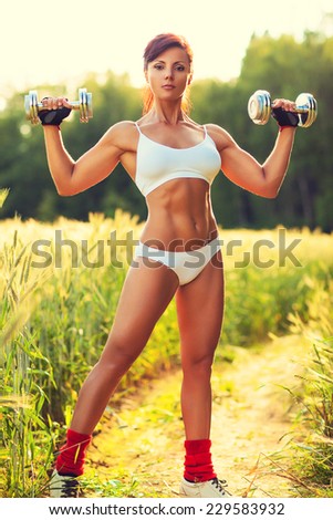 Young sexy woman in white clothing sports training at summer field. Chrome heavy dumbbells in hands. Res sunset light.