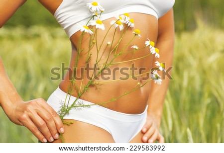 Young sports woman tummy with camomile flowers. White clothing and nature field background.