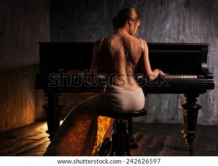 Young elegant woman in evening dress with naked back playing piano in retro style interior. Dark colors and backside view.