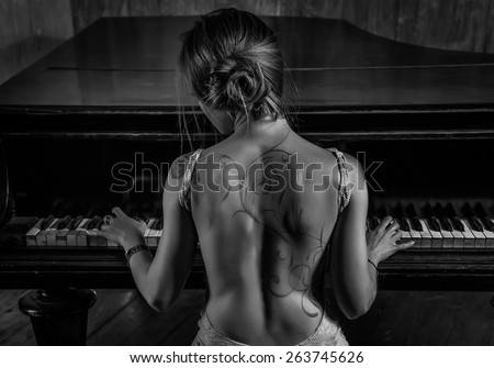 Young elegant woman in evening dress with naked back playing piano. Tattoo on back and black and white dark colors.