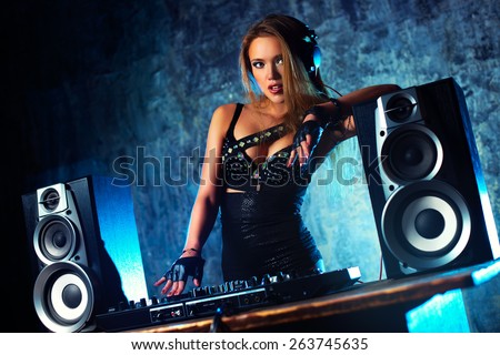 Young sexy woman dj playing music. Big loud speakers, headphones and dj mixer on table. Camera angle view.