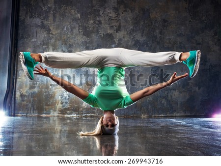 Young woman hip-hop dancer standing on head on wall background.