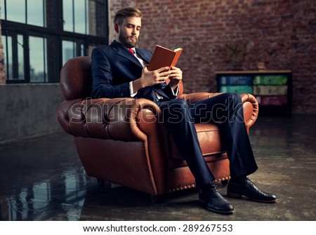 Young handsome businessman with beard in black suit sitting on chair reading book. Focus on face.
