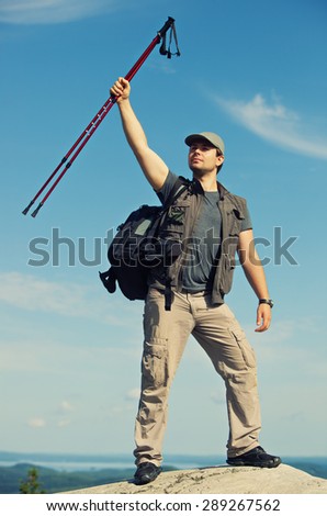 Young man tourist with backpack standing on mountain top and holding sticks above head.