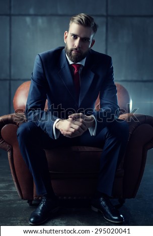 Young handsome businessman with beard in black suit sitting on chair and listens attentively to speaker.