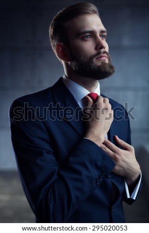 Young handsome man with beard in black suit straightens his tie and looking aside.