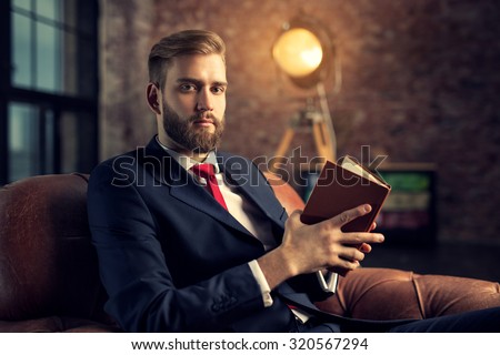 Young handsome businessman with beard in black suit sitting on chair reading book.