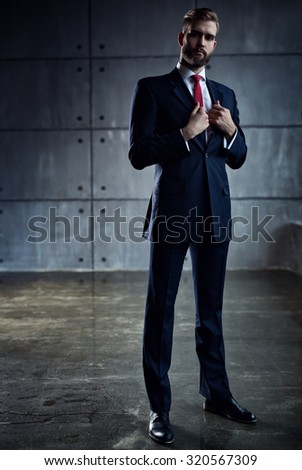 Young handsome businessman with beard in black suit standing on wall background.