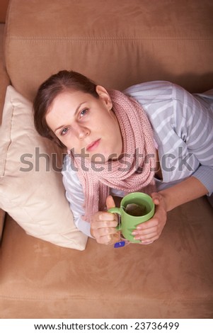 A young woman is sick. She is lying on the couch and is sneezing. She has a tea in her hand.