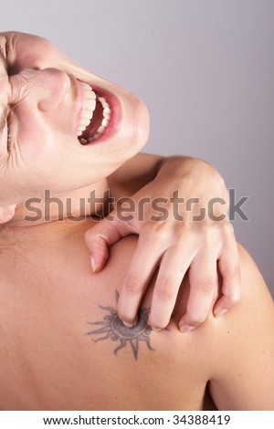 Young woman with sever back pain. She is holding her schoulder. Over grey background. She has a tattoo on her shoulder.