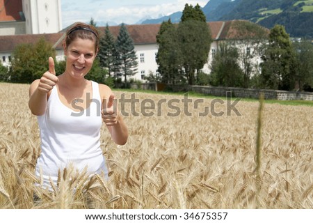 A young woman gives a thumbs up sign in the yellow field under blue sky in a rural alpine setting. Village in the background. Focus is on the woman.