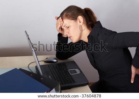 Young woman sitting at her laptop with a lot of work in front of her. She has a pain in her neck / back.