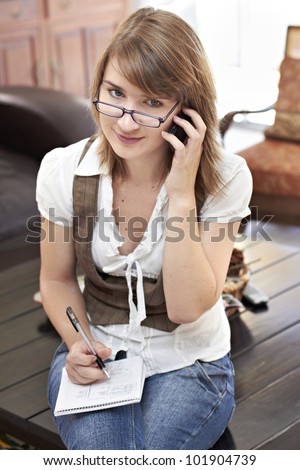 Young lady sitting on table chatting on cellphone while writing on paper pad