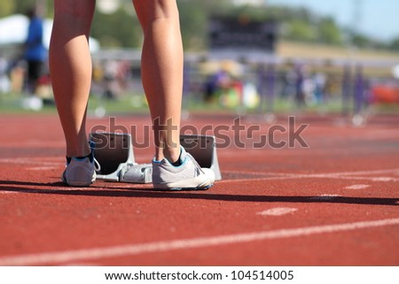 Female athlete sprinter standing behind starting block at the starting line, about to compete in the 100 meter hurdle track & field event.