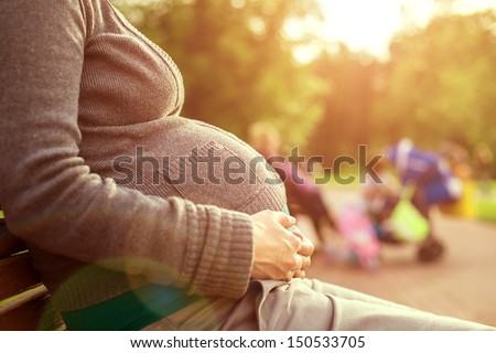 Pregnant Woman Sitting On A Bench. On Background The Children Play. Warm Weather