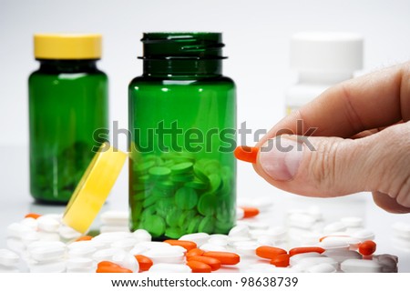 person holding one pill with a variety of pills on table and medicine bottles in background