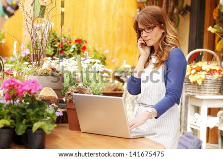 Smiling Mature Woman Florist Small Business Flower Shop Owner. She Is Using Her Telephone And Laptop To Take Orders For Her Store. Shallow Focus.