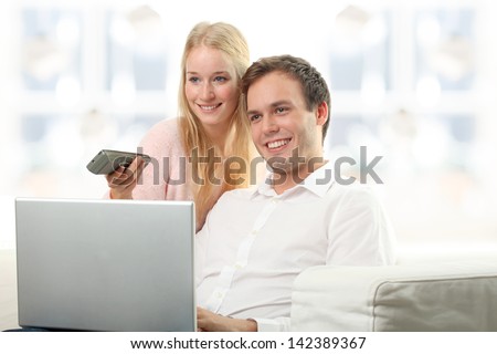 Young serene couple laughing together in their living room at home. Young woman turn on tv while her husband working on laptop.