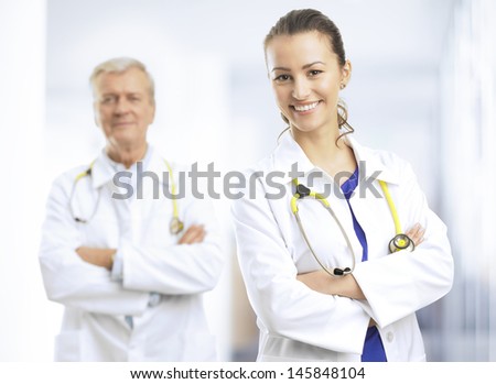 Smiling young female doctor standing with arms crossed in hospital ward Her colleague in the background Smiling young female doctor standing with arms crossed in hospital ward Her colleague in the background