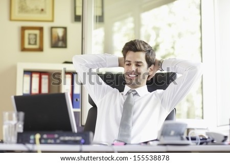 Portrait of a relaxed young businessman smiling and sitting in an office chair with hands behind head. Horizontal shot, Shallow focus.