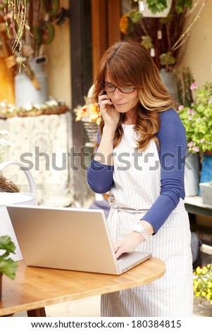 Small flower shop owner working on laptop in her shop and making call.