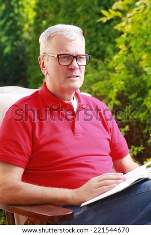 Portrait of old man sitting at garden at home and reading book.