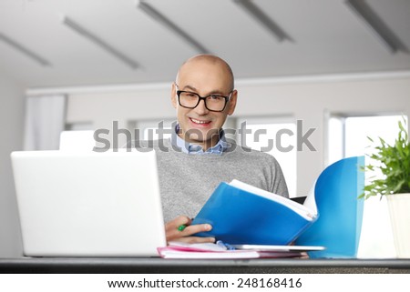 Portrait of casual accountant holding files in hands and analyzing financial data, while sitting at office in front of laptop.