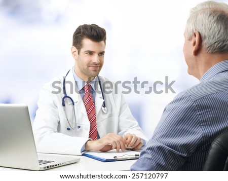 Portrait of young doctor sitting at desk with elderly patient while check up and write diagnosis.