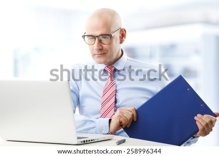 Portrait of executive sales man sitting in front of computer while holding file in his hand. Businessman working at office.