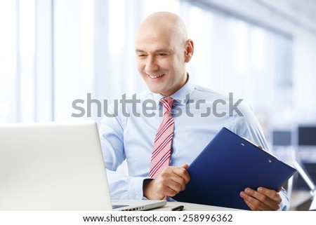 Portrait of busy financial assistant sitting in front of computer while holding file in his hand. Businessman working at office.