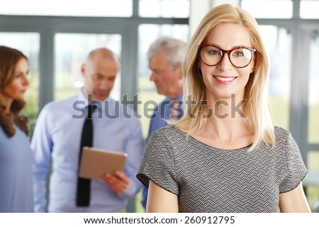 Portrait of executive business woman standing at meeting while her colleagues consulting at background. Business people.
