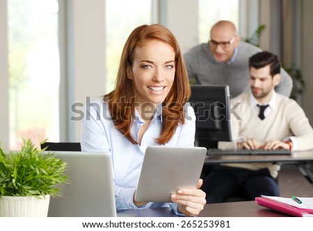 Portrait of attractive business woman sitting at desk in front of computer and analyzing financial data on digital tablet. Business team working at background.