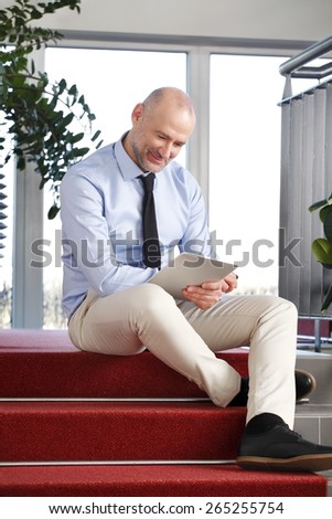 Full length portrait of businessman sitting at staircase and reading his digital tablet.
