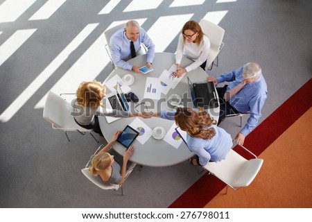 Image from above of business people sitting around conference desk and consulting. Businesswomen and businessmen using laptop and digital tablet while working on business plan.