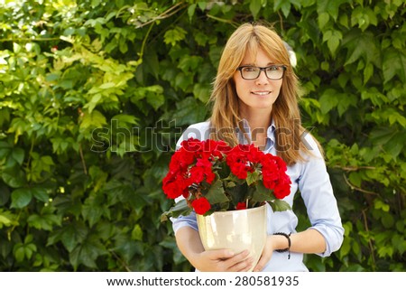 Portrait of attractive woman standing at garden at home while looking at camera and holding potted flower in her hands.