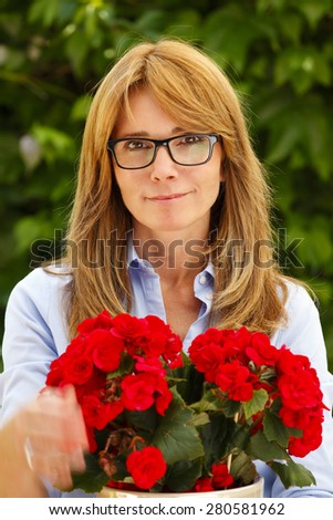 Close-up portrait of attractive woman standing at garden at home while looking at camera and holding potted flower in her hands.