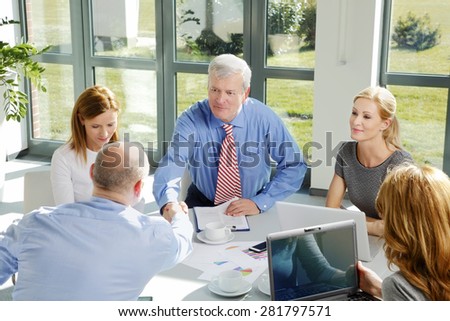Business team sitting around conference table and consulting. Two men shake hands across a white table while three businesswomen smile beside them and working with laptop.