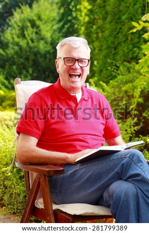 Image of senior man sitting at nursing home garden and reading book while looking at camera and laughing.